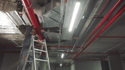 An employee who does the installation of ventilation systems in a new house. The master stands on a ladder and carries out installation of gas pipes and networks fixed on a ceiling.