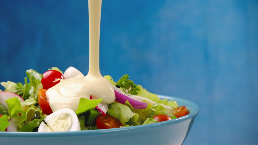 Mayonnaise sauce pouring onto vegetable salad, healthy and dietary food from fresh ingredients with salad dressing in bowl on blue background, slow motion | Shutterstock HD Video #1062107641