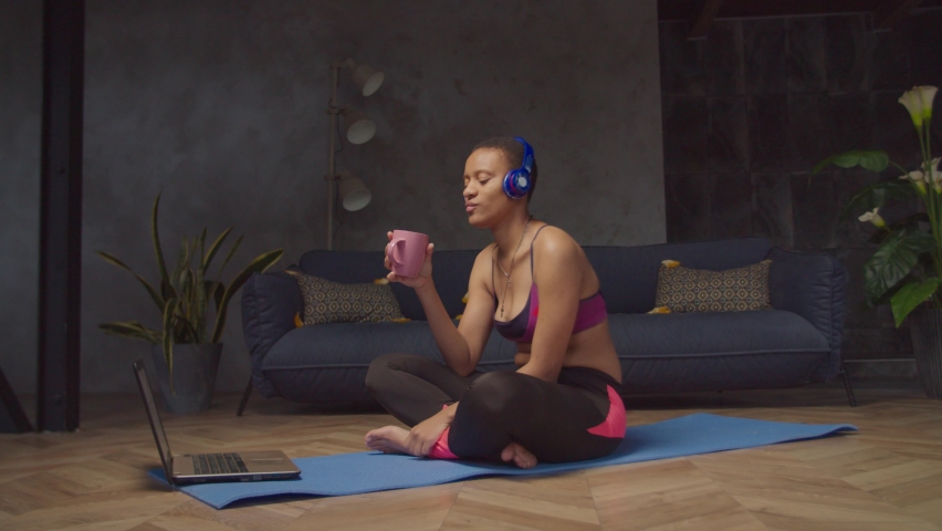 Positive carefree african woman in sportswear sitting cross legged on exercise mat, listening to music in wireless headphones and choosing playlist on laptop while relaxing after training at home.