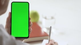 Close-up shot of green screen template smartphone in female hands at home offise, girl is watcing content, makes notes in a notebook. Modern technology and information concept.