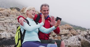 Senior hiker couple with backpacks sitting on the rocks and waving while having a video call on smartphone while hiking near sea shore. trekking, hiking, nature, activity, exploration, adventure