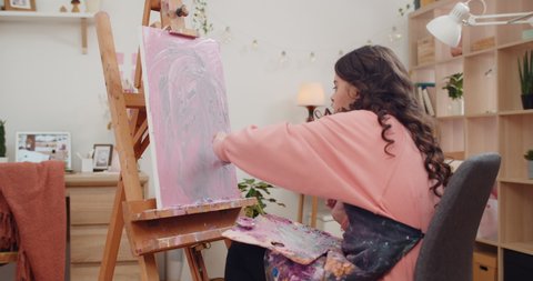 Long haired brunnette girl painting modern picture while sitting in front of molbert. Talanted teenager creating while applying paint and doing strokes on canva. at home. Concept of art