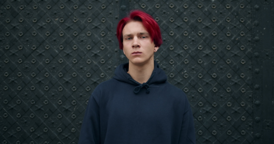 Portrait of millennial male hipster crossing arms and looking to camera. Crop view of young angry guy with dyed red hair and earring standing outdoors. Concept of people and emotions