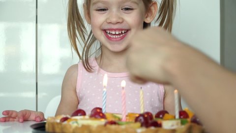 Close up of little girl smiles, laughs and blows out candles on festive cake with fruits. Happy child celebrates birthday. Holiday concept