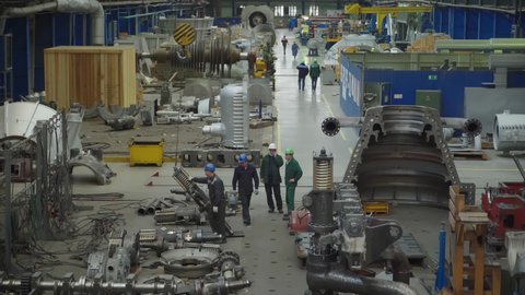 A huge manufacturing facility room. The workers in protective gear are preparing instruments for a new steam turbine to be built. Moving heavy equipment with cranes. Metal parts. Power engineering.