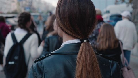 Back view of young stylish woman in a leather jacket walking in the downtown, in crossing the road through the crowd. Footage of blurred people coming in downtown from the back, busy city life 4k