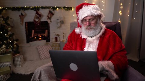 Modern Santa Claus. Cheerful Santa Claus working on laptop and smiling while sitting at his chair with fireplace and Christmas Tree in the background.