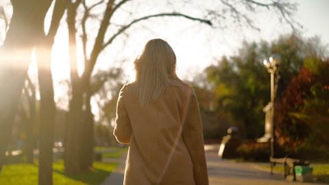 Unrecognizable Beautiful Business Woman in a brown coat walks through city park to work in the office in autumn in sunset light. Back view. Slow motion