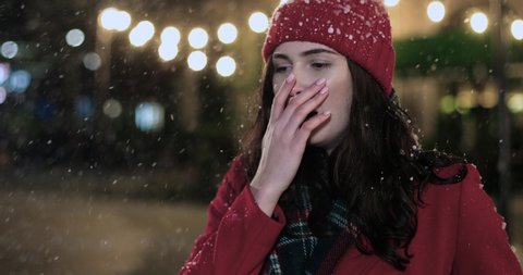 Close up of unhappy sick Caucasian woman in red coat and hat outdoors coughing badly while standing on street alone in evening. Unwell female having virus infection. Christmas season. Holidays concept