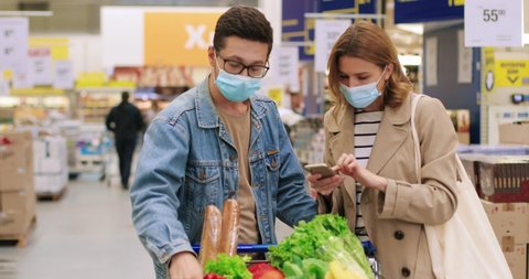 Portrait of Caucasian happy young family in masks in supermarket checking shopping list on cellphone with food products in shopping cart. Woman tapping on smartphone while buying food Portrait concept