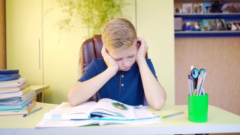 portrait of a tired young blond boy who teaches lessons with a book and notebook at the table in the room. Teen schoolboy falls asleep and sleeps while studying. Close up face of an exhausted young 