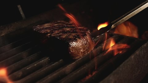 Juicy beef steak is fried on fire coals on iron grill on dark background in flame of smoke. Barbecue in evening, chef turns meat on grill with tongs. cooking dinner in a hot flame.