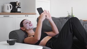 Young millennial woman at home applying cloth mask on face and doing video call. Wellness relaxing time 