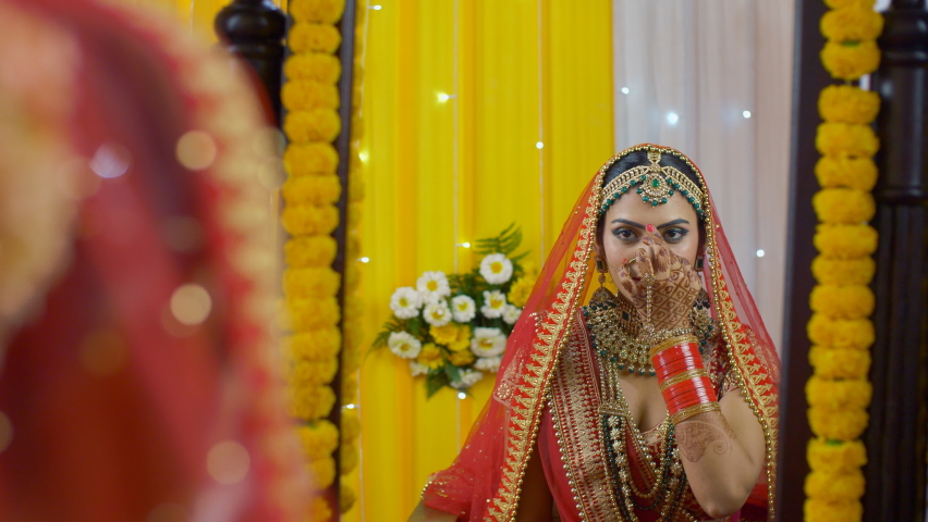 Lovely Punjabi bride putting on Bindi while looking in the mirror - bridal look. Attractive young girl in red-gold bridal Lehenga and heavy jewelry - Indian wedding preparations