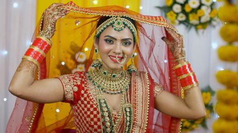 Beautiful Indian bride wearing red Lehenga with Chuda for her wedding. Medium shot of a cheerful and smiling bride slowly lifting her ghoonghat or veil - Hindu wedding