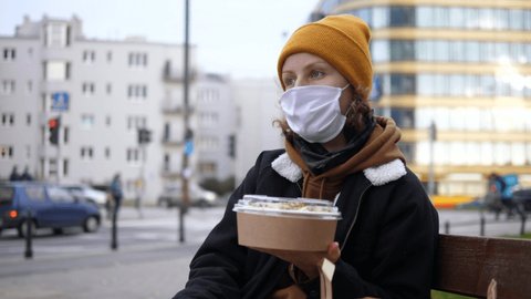 White woman takes off mask to eat green salad in single use recyclable package on street. Healthy vegan fast food business during covid-19 outbreak