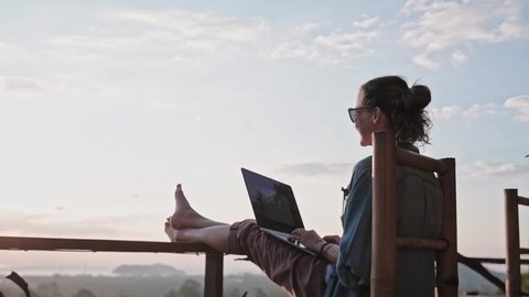 Freelancer woman sits with a laptop on a high bar stool. Amazing sunset views and dense jungle by the blue sea. A nice girl with glasses and a ponytail works on a computer in a species cafe.