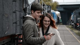 A guy and a girl are standing on the platform of the railway station and together watching a video on the phone