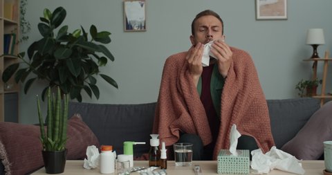 Adult ill guy talking paper handkerchief and sneezing while sitting on sofa covered with blanket.Young bearded man feeling bad while having flu or cold and taking cure at home.