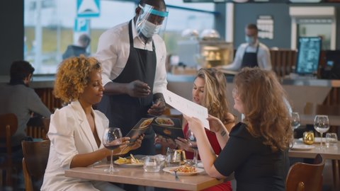 African waiter in safety mask and shield bringing menu to young women in restaurant. Three multiethnic female friends hanging out together in modern restaurant