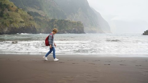 4K Teenage boy with red backpack walking by black sandy beach stopping looking at ocean waves. Holidays, environment, adolescence, growing up concept.