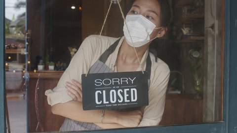 Young Asia girl wear face mask turning a sign from open to closed sign on glass door cafe after coronavirus lockdown quarantine. Owner small business, food and drink, business financial crisis concept