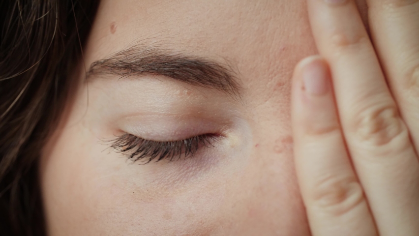 Close up of Woman covering half her face with hand and opening other blue eye. Young female model with natural make-up and long eyelashes looking at camera. Slow-motion, macro extreme close-up, 4K.