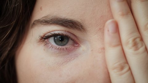 Close up of Woman covering half her face with hand and opening other blue eye. Young female model with natural make-up and long eyelashes looking at camera. Slow-motion, macro extreme close-up, 4K.