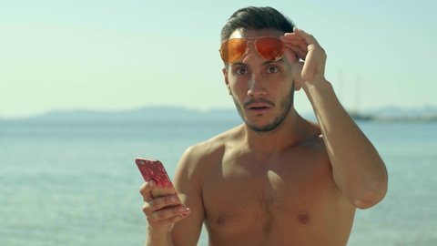 The young man in sunglasses, sunbathing by the sea and spending time on his mobile phone, looks at the camera in surprise, sees a news on the phone.