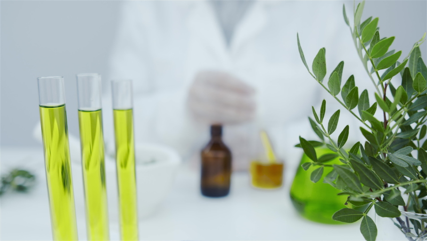 Oil drips from pipette into bottle. Pipette in hands of laboratory assistant. Cosmetics oils based on natural ingredients. Mortar with press, branch of plant, glasses with natural oil on white table. Royalty-Free Stock Footage #1062192598