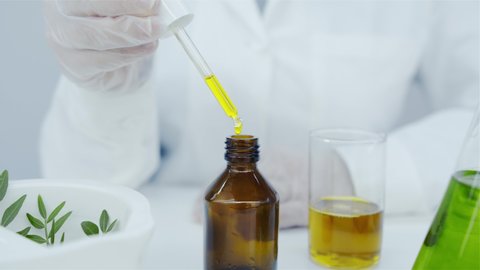 Oil drips from pipette into bottle. Pipette in hands of laboratory assistant. Cosmetics oils based on natural ingredients. Mortar with press, branch of plant, glasses with natural oil on white table.