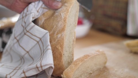 A slice of white bread being cut from a bread loaf with a knife on a wooden cutting board while another slice of bread is laying beside. Slow motion with shallow depth of field.