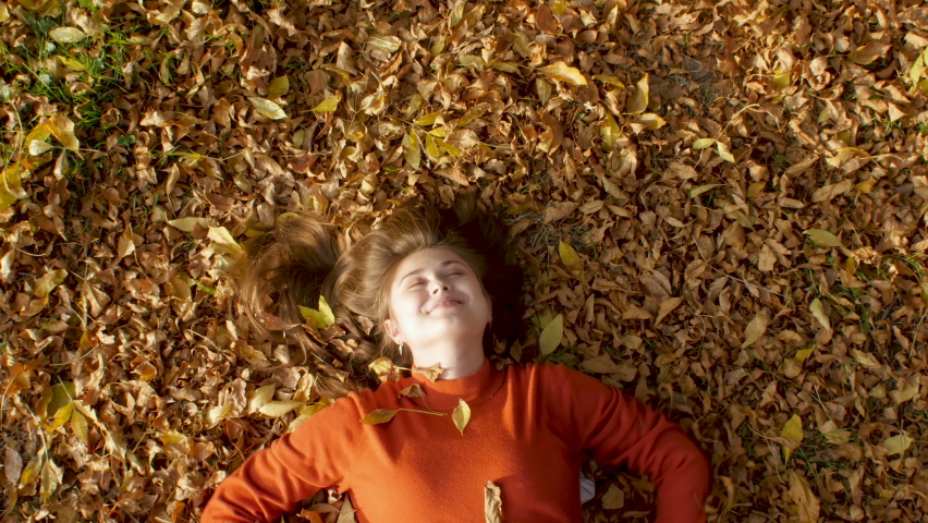 cheerful beautiful young girl lying in autumn foliage and enjoying life. Happy woman playing throws up dry leaves in park, top view