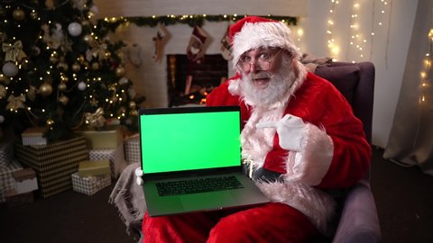 Santa Claus in glasses sitting in his rocker near christmas tree. holds a laptop with green screen and attracts attention to it. Christmas spirit, holidays and celebrations concept 4k footage