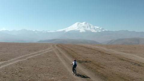 Woman riding on electro e-bike bicycle at mountain plain on snowy peak and valley landscape from drone. Traveler woman bicycling in mountain bike on highlands on snowy Elbrus Dzhily Su landscape.