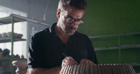 Low angle of focused male artisan using tool to remove clay and create ornament on clay bowl in workshop