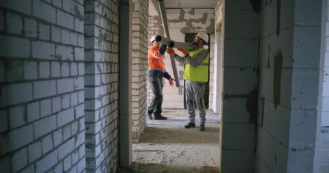 Tracking shot of male contractor taking pipes from colleague and walking in brick hallway during work on construction site