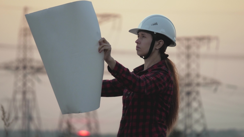 Silhouette of an engineer looks at the project of construction of a high-voltage power plant. The engineer is reading the drawing. engineer works in the field next to the electricity pylons at sunset.