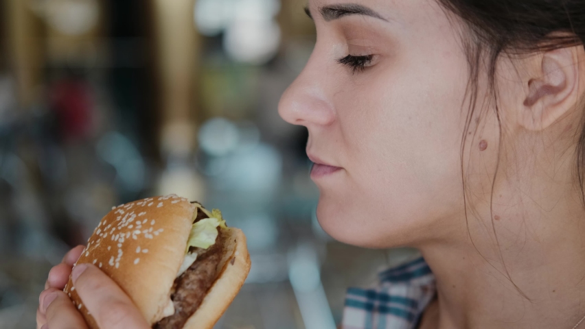 Young hungry woman bites a burger in a fast food restaurant. Girl eating a hamburger close up. | Shutterstock HD Video #1062202462