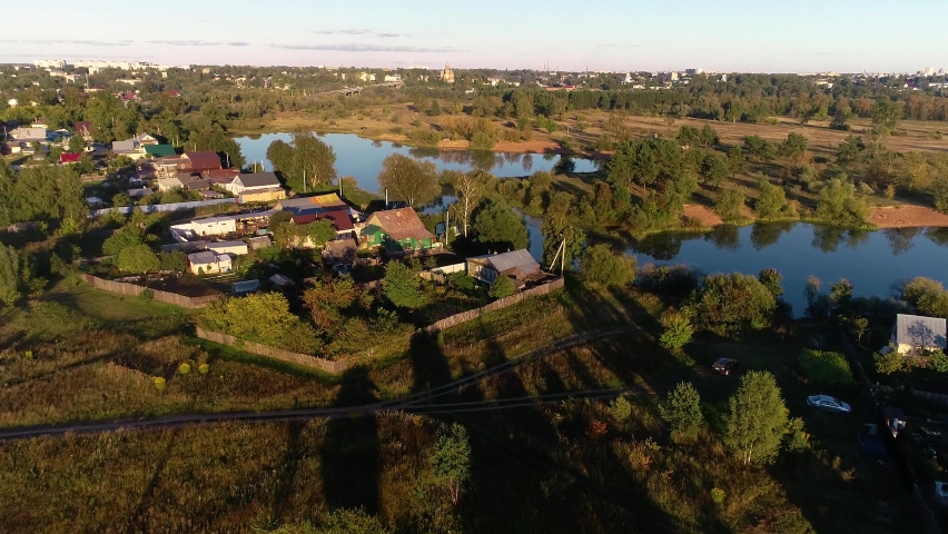 The lake shore, beautifully lit by the evening sun. Wooden rural houses are visible. The open spaces are colored orange. Long shadows fall from trees and bushes. The city was visible in the distance.
