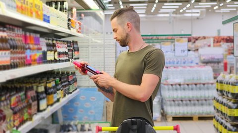 Young man customer is choosing beer in alcohol department of grocery shop. He taking bottle off the rack, comparing trademarks, prices, reading labels. Close up, slow motion