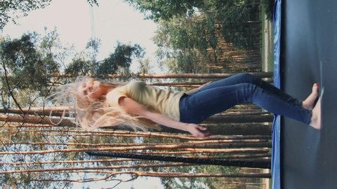 Woman in light summer shirt and jeans looking at camera and smiling jumping on large trampoline with protective barrier with tall trees around. Video with Vertical Screen Orientation 9:16