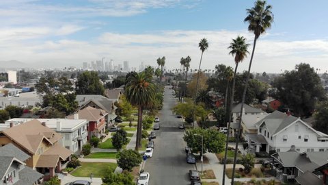 Aerial slow flyover street in Los Angeles neighborhood between palm trees with downtown Los Angeles in the distant background, sunny day, California