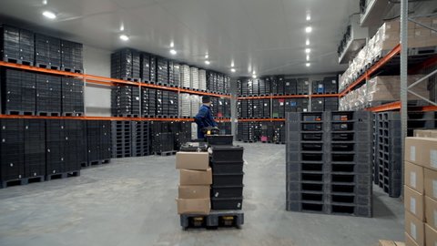 Warehouse at the Modern Factory. A worker delivers finished products on a forklift and places them on shelves in a warehouse. Cardboard boxes, plastic boxes. Stock. Storage room. Wide shot