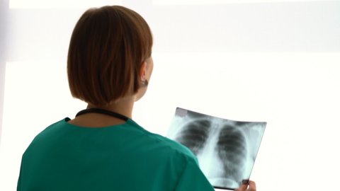 Young woman doctor is looking at chest x-ray while standing in modern clinic. Back view of female specialist examining picture and holding it in her hands during working day in hospital. 