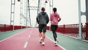 Back view of couple in sportswear running on bridge with cloudy sky, slow motion