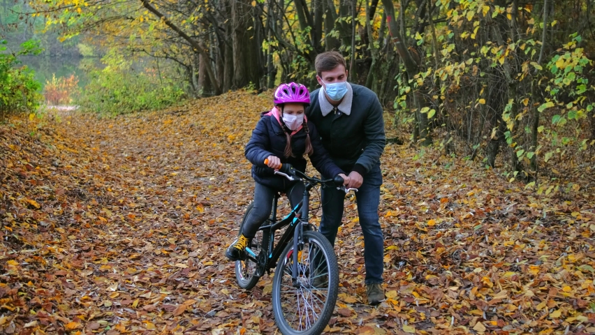 A young father teaches his daughter to ride a Bicycle in a protective helmet. they have medical masks on their faces. walking during the covid-19 coronavirus pandemic. | Shutterstock HD Video #1062223267