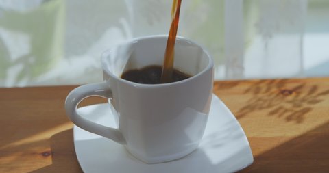 Man's hand pours coffee into a cup on a wooden table by the window. Drink fresh coffee in the morning at home. Man pours hot aromatic coffee into a white cup from a coffee machine in the room. 