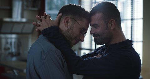 Cinematic shot of young happy smiling married homosexual male gay couple is enjoying time together is hugging and kissing as sign of timeless love in a kitchen at home.