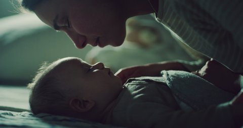 Cinematic shot of neo mother is caressing and kissing with affection her newborn baby while is sleeping peacefully with sweet dreams in a crib with soft blanket in a nursery at night.